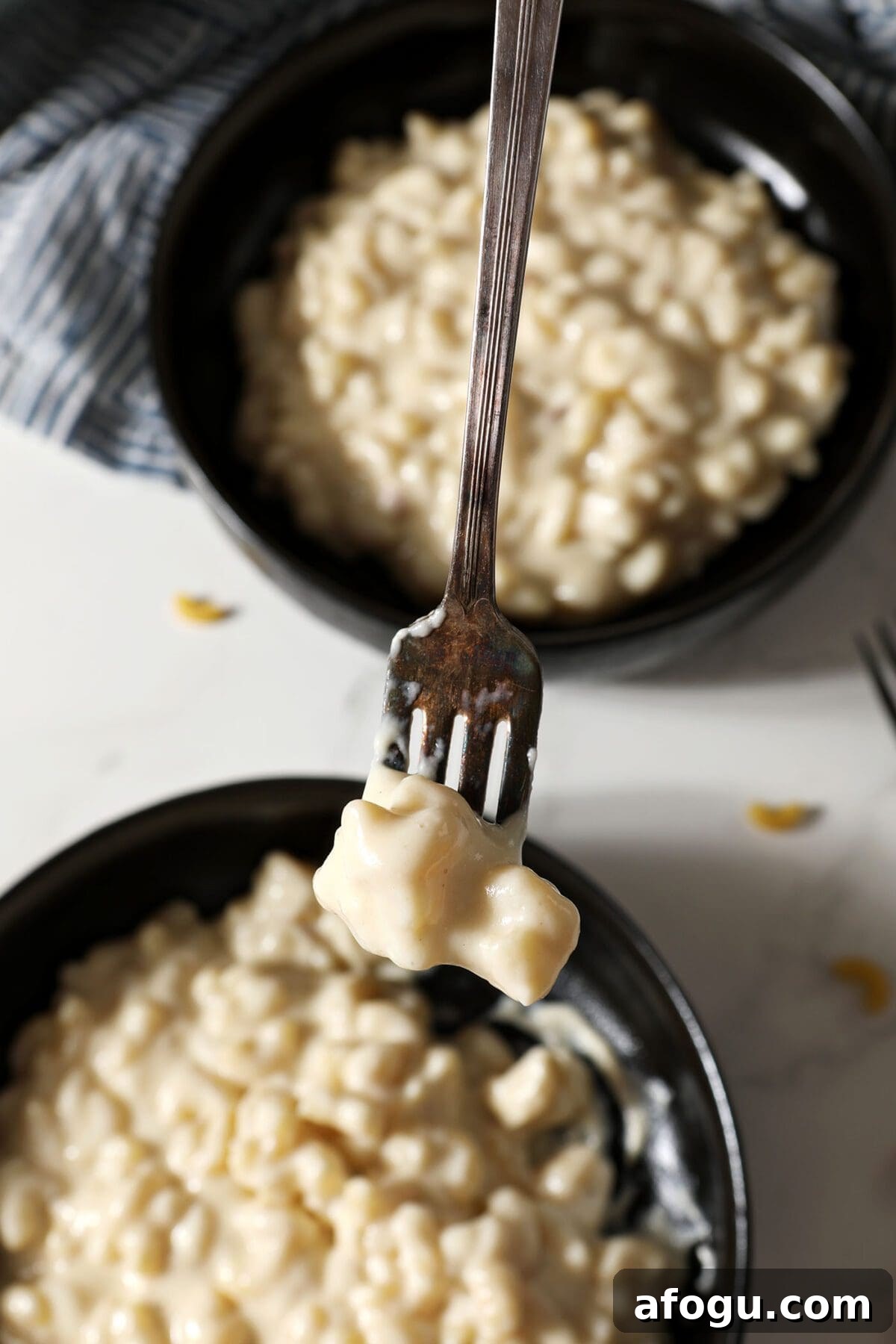 A fork holds a bite of three cheese Mac and cheese above two black bowls holding more macaroni and cheese