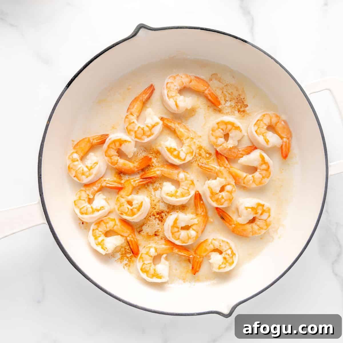 Overhead view of shrimp cooking in a white skillet on a marble surface.