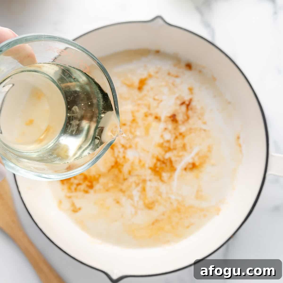 Pouring liquid from a measuring cup into a white pan with browned bits on the bottom; a wooden spoon lies beside the pan on a marble surface.
