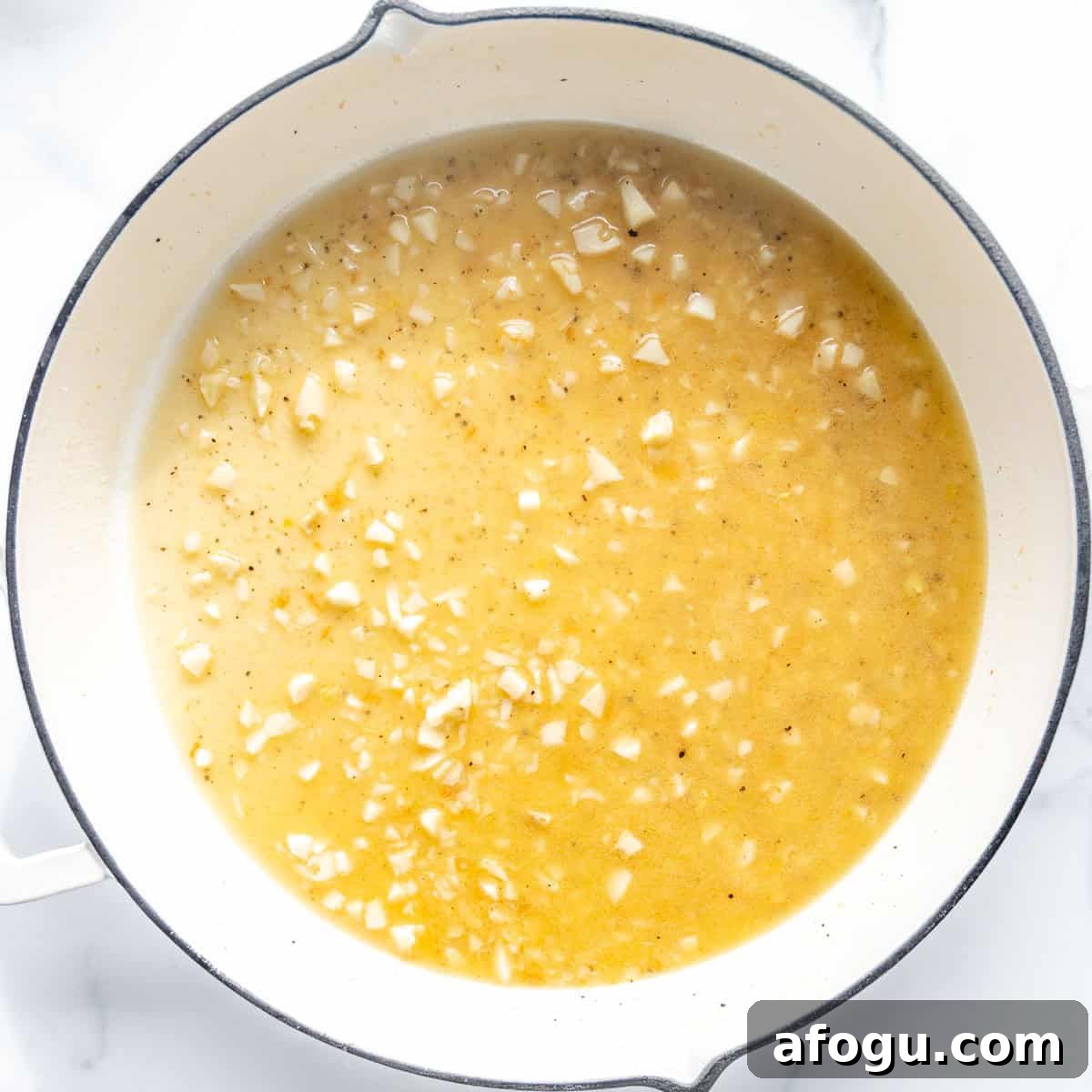 A white pot filled with chopped garlic in a light brown liquid mixture, possibly a sauce or broth, on a marble surface.