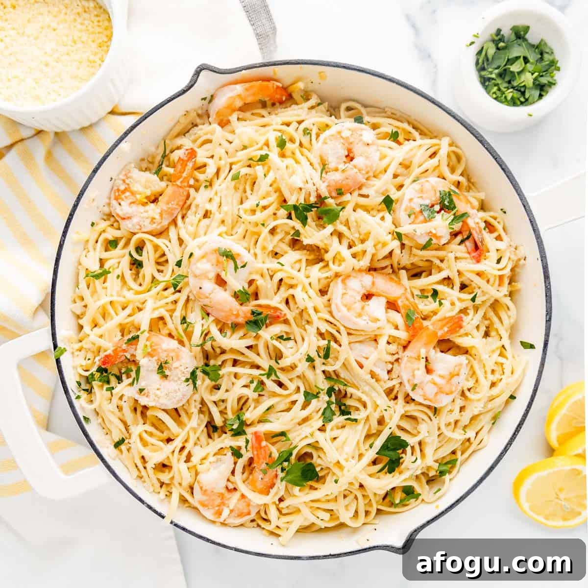 Shrimp scampi pasta in a white pan, garnished with parsley. Nearby are lemon slices, a small dish of grated cheese, and a bowl of chopped herbs on a striped cloth.