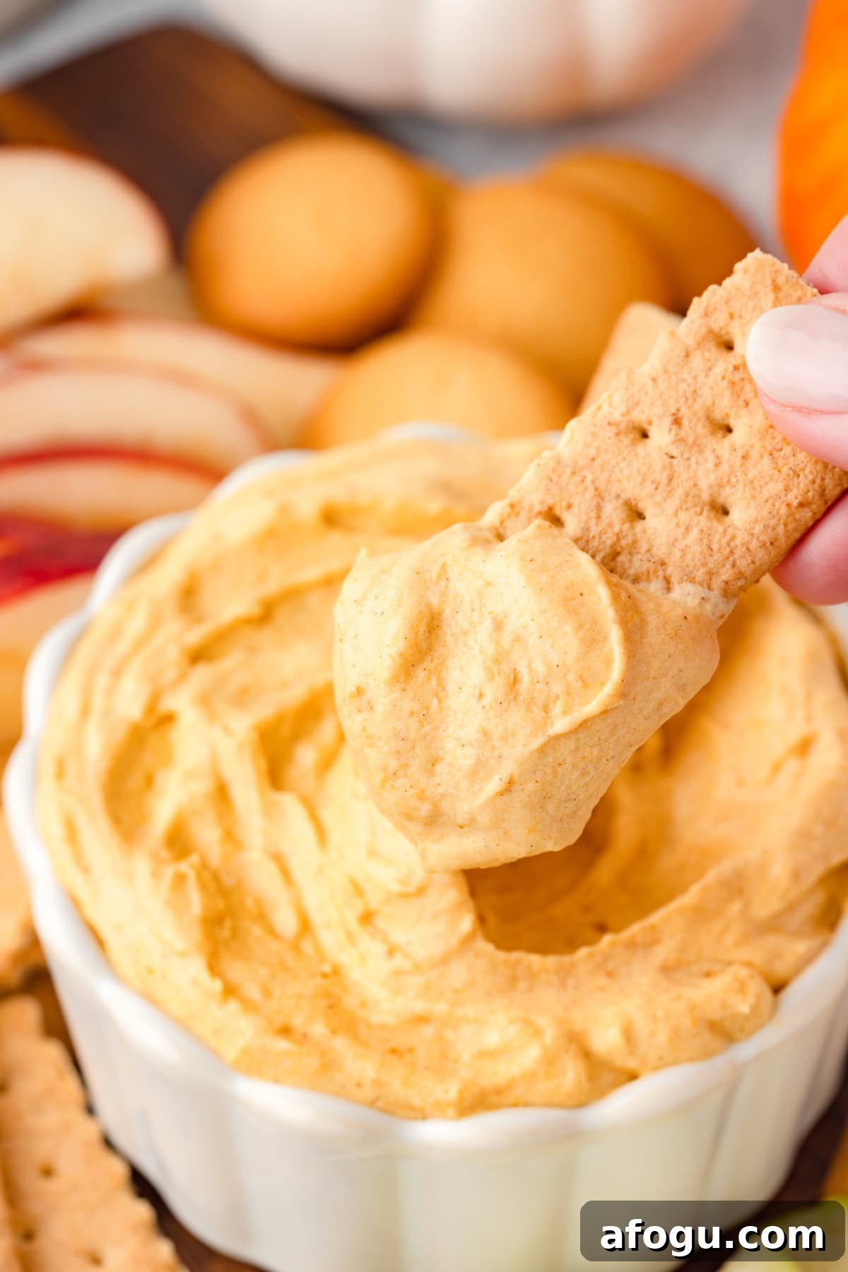 A hand dips a graham cracker into a bowl of creamy pumpkin dip.