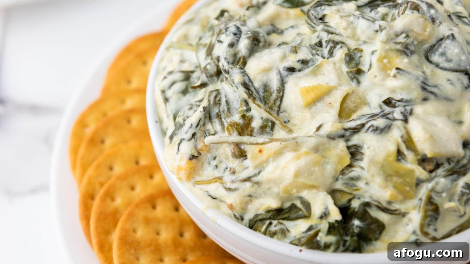 A bowl of creamy spinach artichoke dip with a side of round crackers on a white surface.