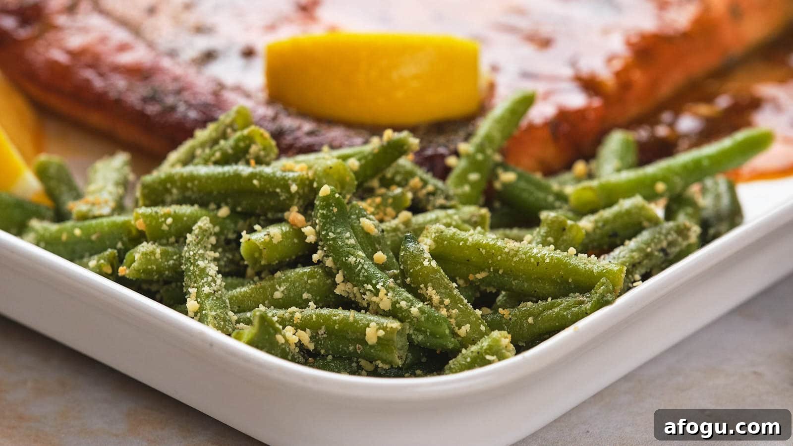 A plate with green beans sprinkled with seasoning and grated cheese, alongside a lemon wedge and a portion of glazed salmon.