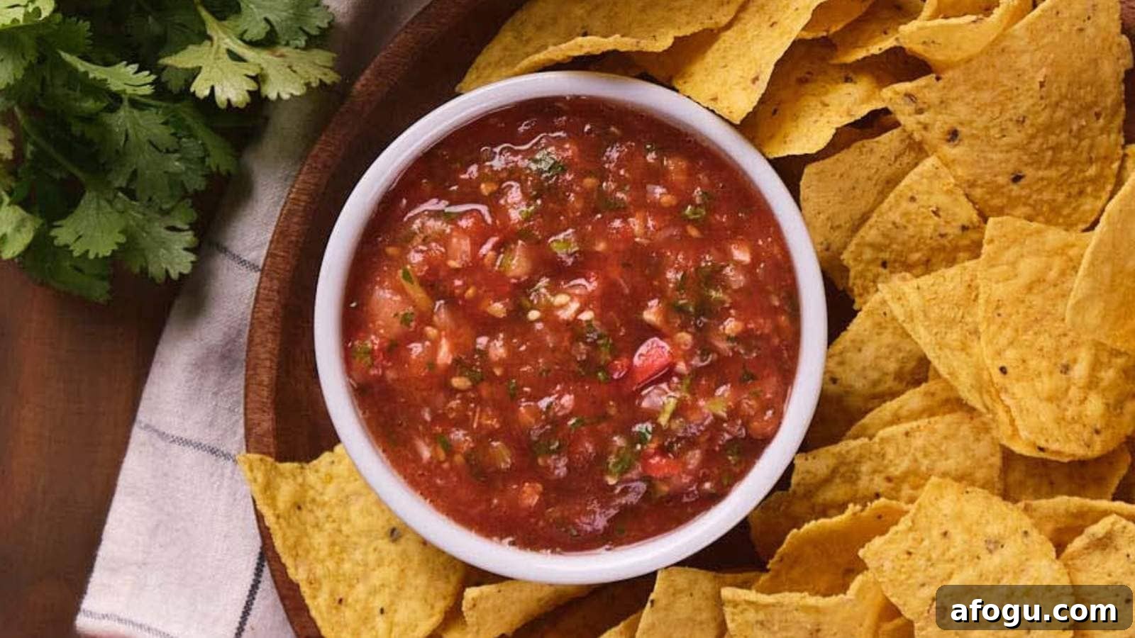 A white bowl of salsa surrounded by tortilla chips on a wooden platter, with a sprig of cilantro nearby.