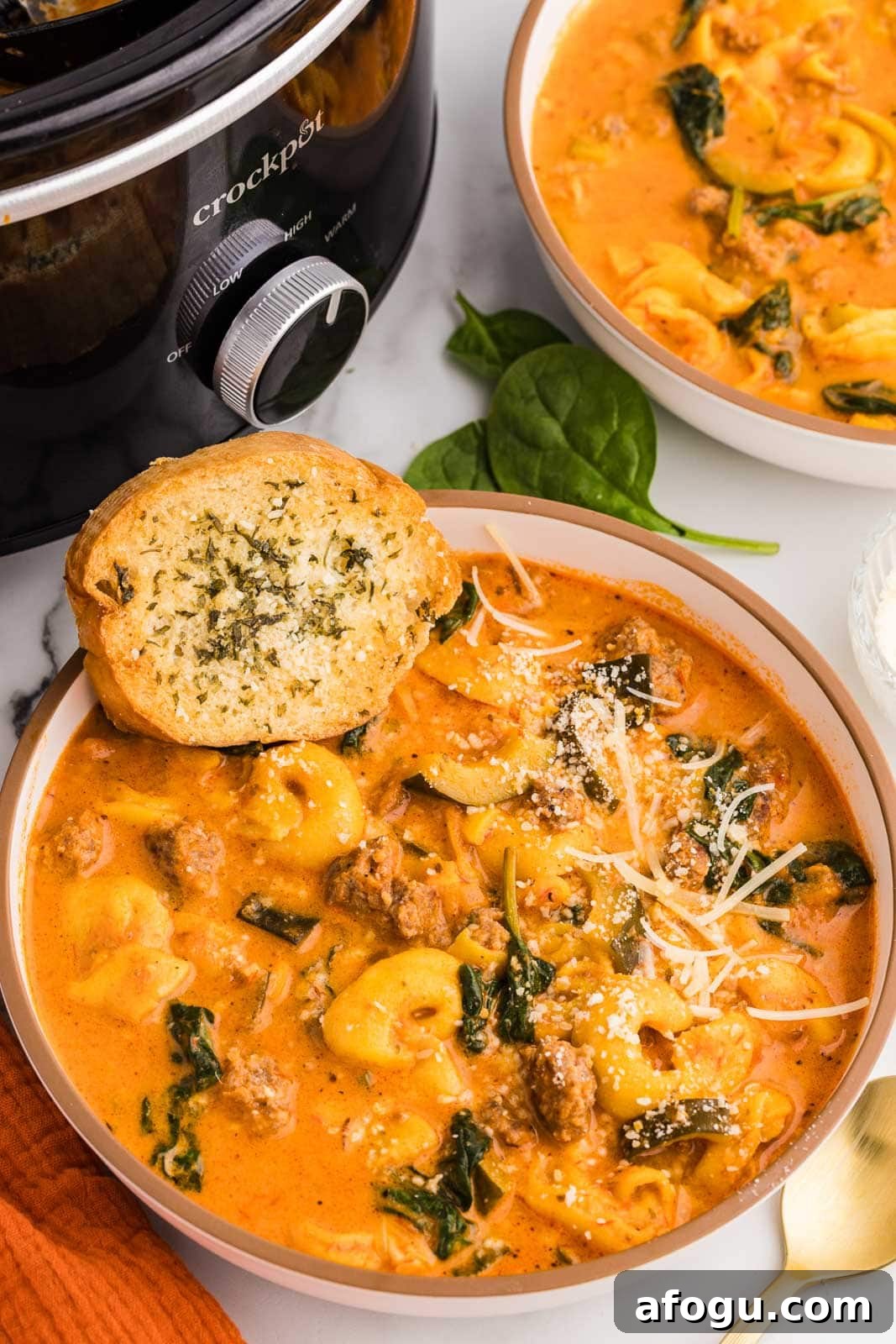 two bowls of tortellini soup, one with garlic bread on side, with black slow cooker in background