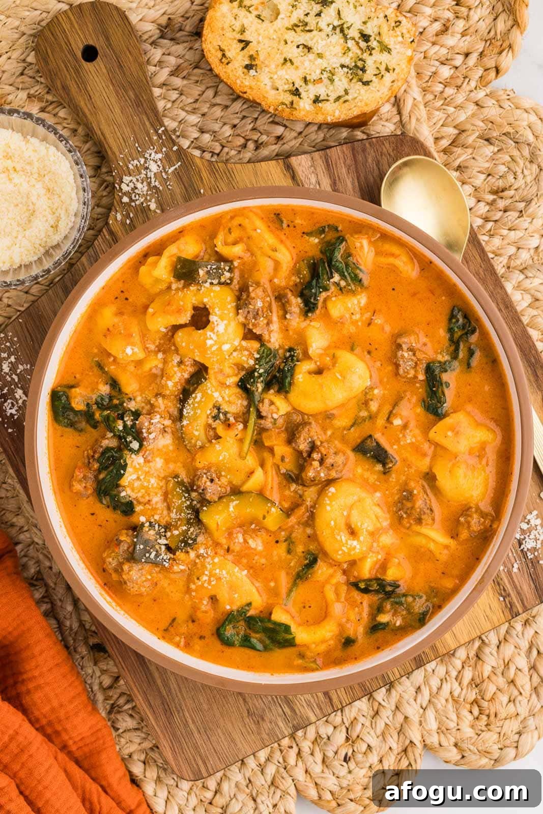 overhead view of bowl of tortellini soup on wooden cutting board and tan placemat, surrounded by gold spoon, bowl of parmesan cheese, and garlic bread