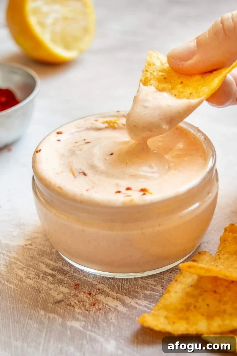 A hand dipping a yellow tortilla chip into a thick crema dip with a slight orange tinge.