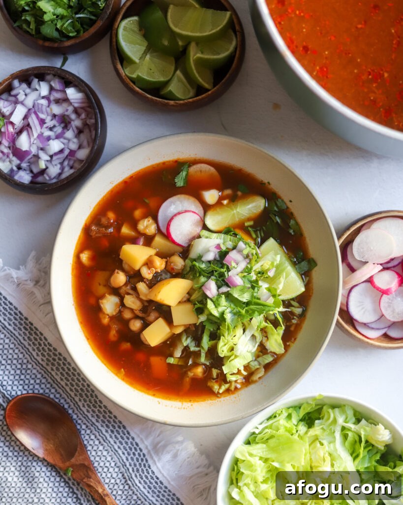 overhead view of a bowl of homemade vegan pozole rojo next to containers of garnishes like sliced lettuce, radishes, onions, and limes, next to the cooking pot.