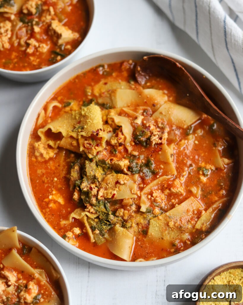 Overhead shot of 3 bowls of Lasagna soup, one large bowl in the middle filled with noodles, tomatoes, kale, and nutritional yeast