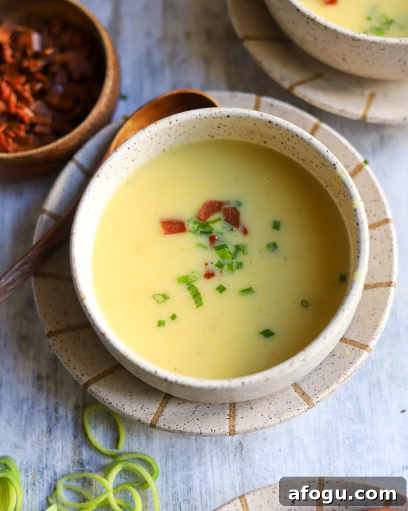 overhead view of a bowl full of creamy potato leek soup with a small bowl of bacon bits and some chopped leeks on the table