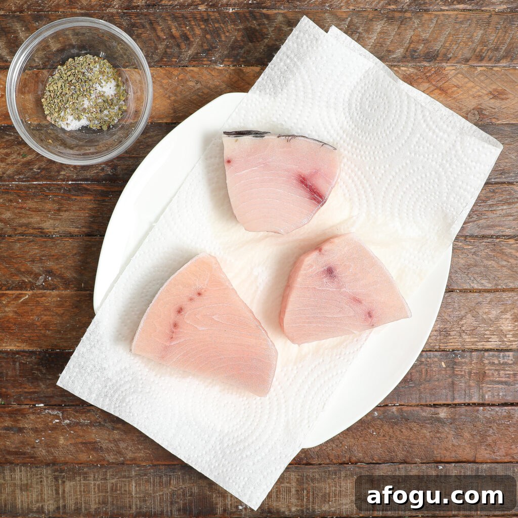 Three raw fish steaks rest on a paper towel on a white plate, ready for Swordfish Piccata, with a small glass bowl of mixed dried herbs and salt nearby, all set on a rustic wooden surface.
