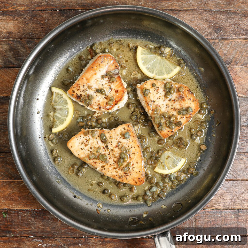 Three pieces of seared Swordfish Piccata in a stainless steel skillet with capers, lemon slices, and a light sauce, set on a rustic wooden surface.