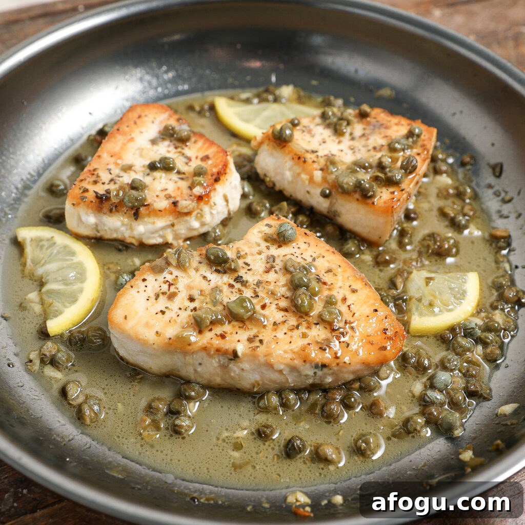 Three Swordfish Piccata fillets seared in a pan with a sauce of capers, lemon slices, and herbs, all simmering together in a light, savory broth.