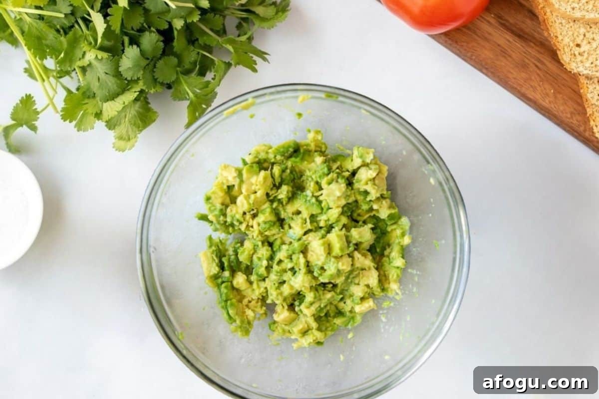Mashed avocado with lime juice and salt in large mixing bowl with bread and tomatoes on cutting board.