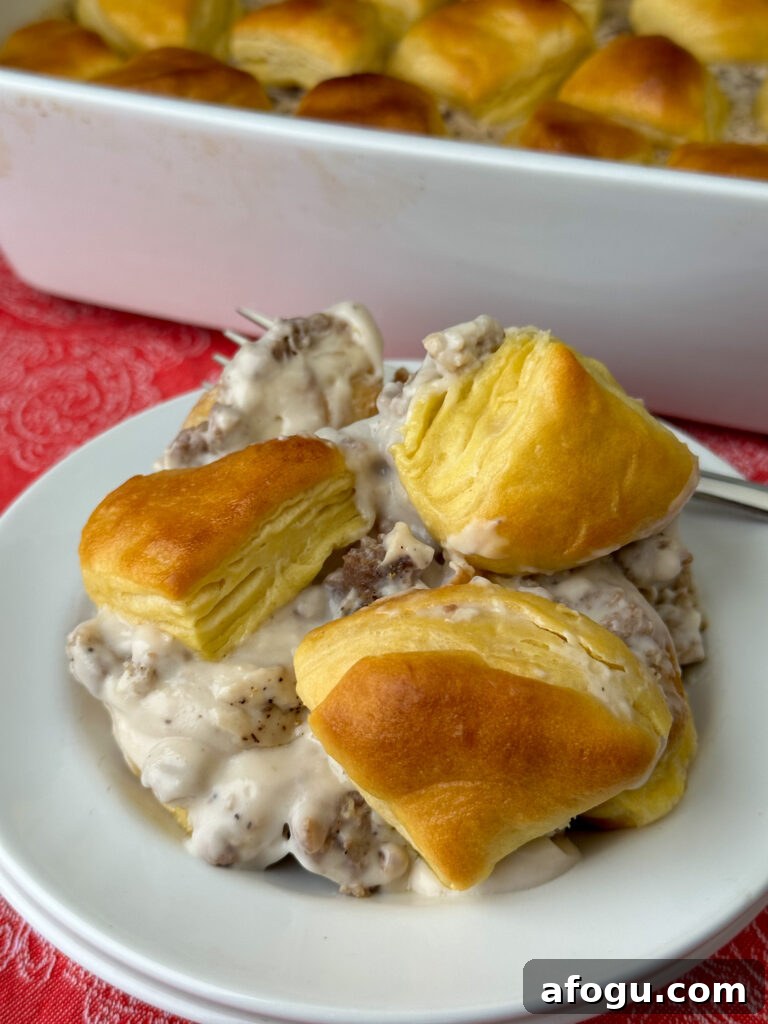 Pieces of golden brown biscuits resting on top of a creamy gravy with extra sausage added on a white plate with a fork and the casserole dish in the background.