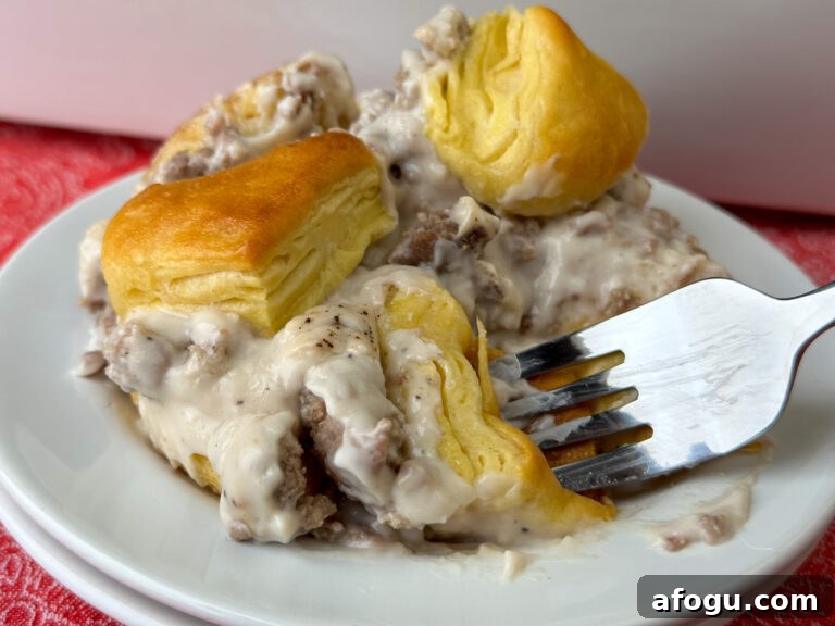 Fork in a warm piece of biscuit with peppered gravy and fresh sausage on a plate full of sausage and gravy biscuit casserole on a red napkin.