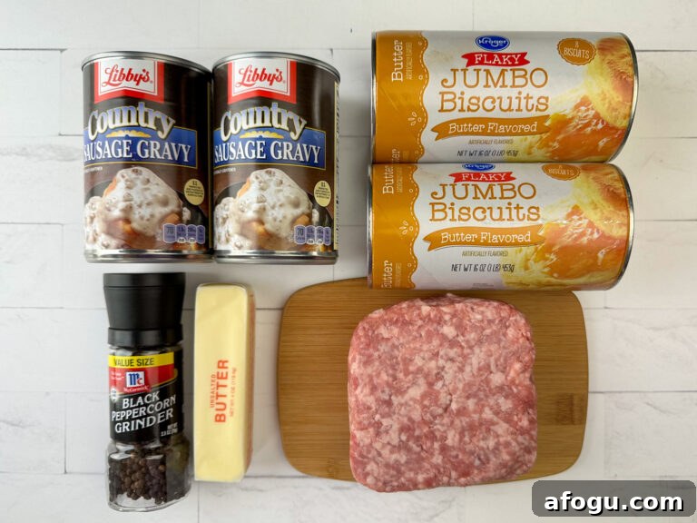 Ingredients to make biscuit and gravy casserole are two cans of country sausage gravy, two cans of flaky jumbo biscuits, black pepper, butter and sausage on a small cutting board all sitting on a white counter top.