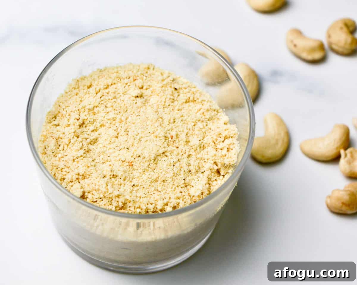 A glass filled with nutty cheese topping on a grey and white marble background with a few cashews sprinkled around.