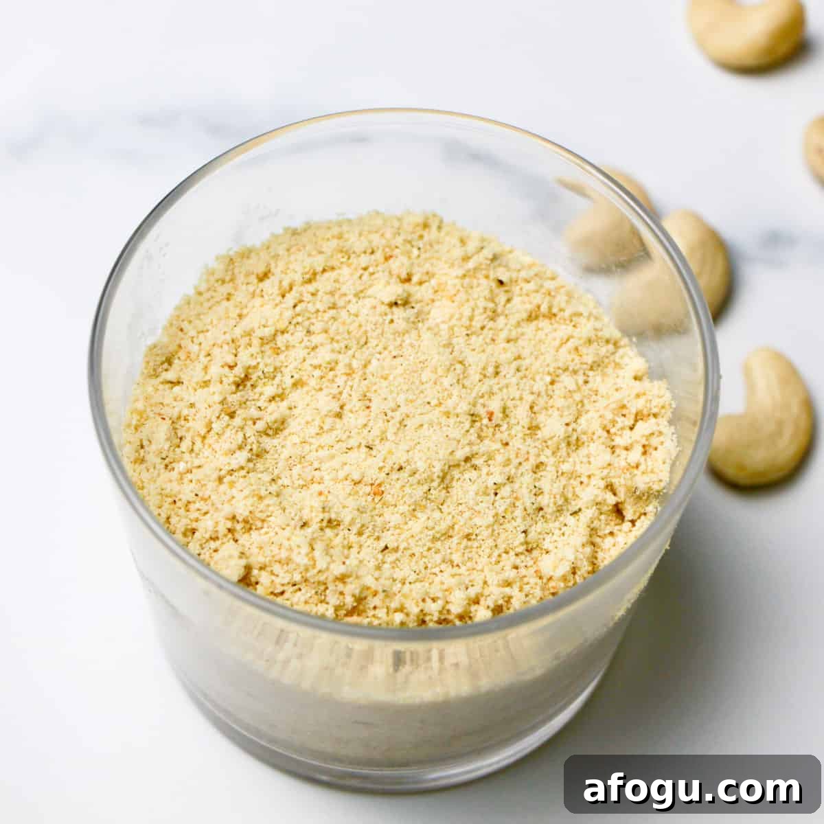 A glass filled with nutty cheese topping on a grey and white marble background with a few cashews sprinkled around.