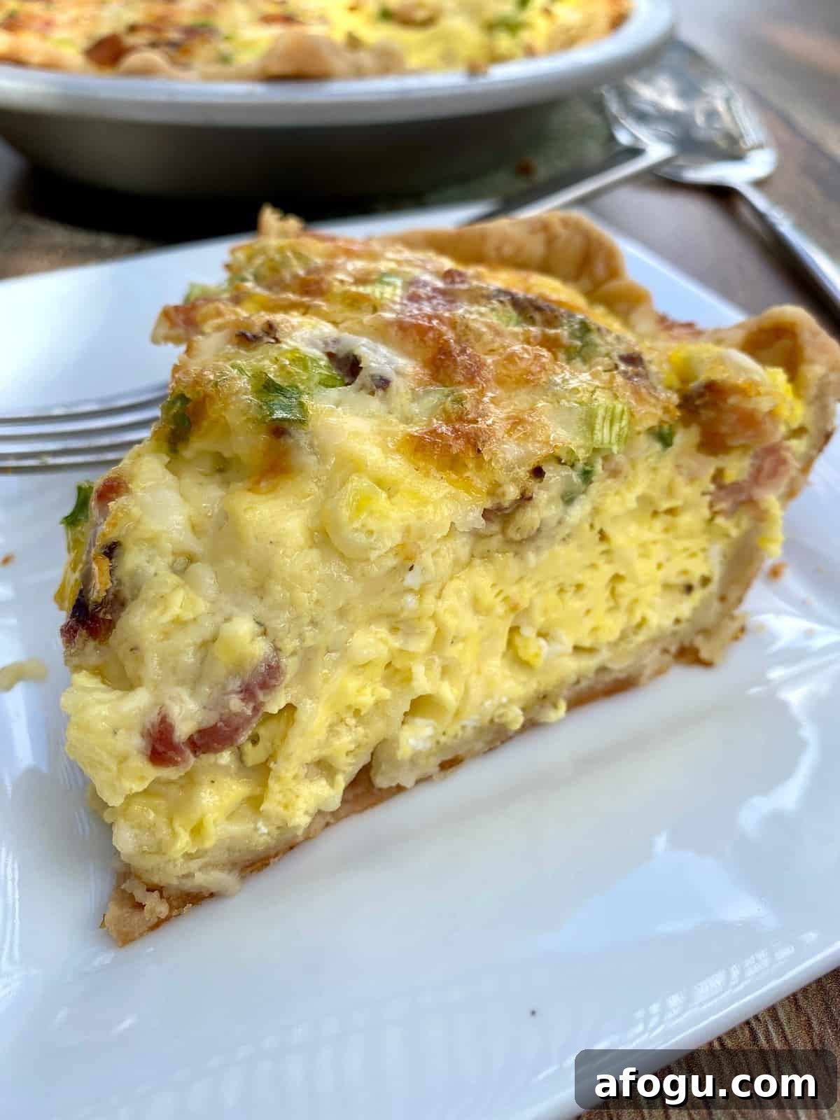 A wedge of quiche on a plate with the quiche dish in the background.